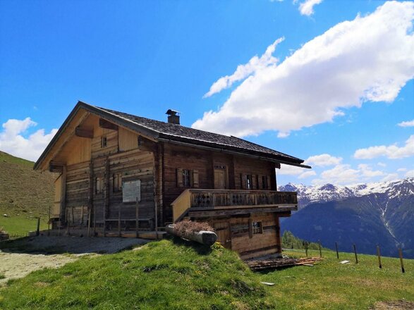 Das Bild zeigt die Schmiedleralm, eine Almhütte aus Holz auf einer grünen Bergwiese. Vor der Hütte befinden sich ein Weg, ein Geländer und einzelne Zaunpfosten. Im Hintergrund sind Berggipfel mit Schneefeldern unter einem blauen Himmel mit weißen Wolken zu sehen.