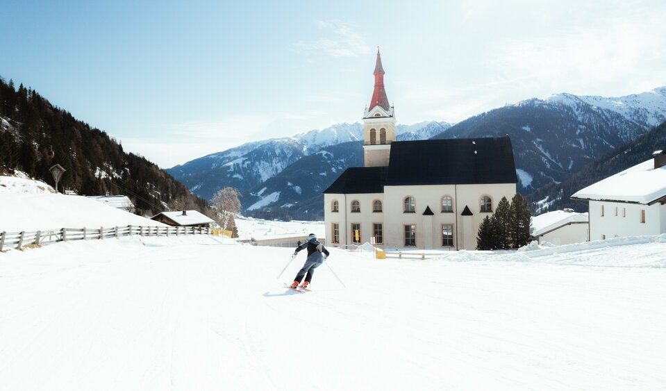 Ein Skifahrer zieht auf einer flachen Piste, die neben der Pfarrkirche vorbei läuft, bei herrlichem Winterwetter sein Schwünge sanft in den Schnee.