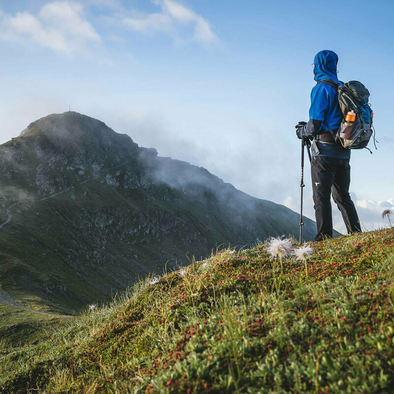 Schöne, frische Morgenstimmung - ein gut eingepackter Wanderer mit langer, dunkelgrauer Berghose, blauem Anorak mit übergezogener Kapuzze und blau-grauem Rucksack und angesteckter heller Trinkflasche blickt zu dem in einiger Entfernung liegenden Bergziel, das sich noch vom morgendlichen, leichten Nebel befreit.