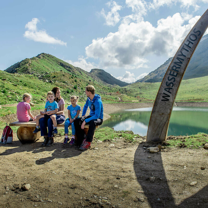 Eine Familie sitzt auf einer Bank am Ufer des Wassermythos Ochsenlacke im Skizentrum St. Jakob i. D..  Im Hintergrund sieht man die dazugehörigen Spielgeräte und das umliegende Bergpanorama, welches durch das schöne Wetter gut zur Geltung kommt.