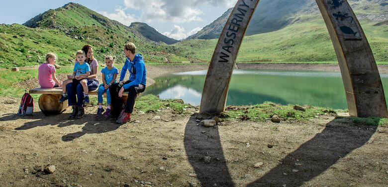 Eine Familie sitzt auf einer Bank am Ufer des Wassermythos Ochsenlacke im Skizentrum St. Jakob i. D..  Im Hintergrund sieht man die dazugehörigen Spielgeräte und das umliegende Bergpanorama, welches durch das schöne Wetter gut zur Geltung kommt.