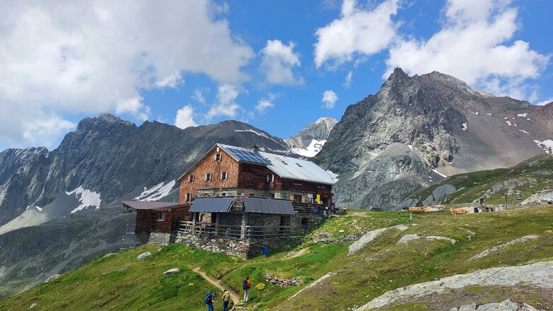 Das Bild zeigt die Bonn-Matreier-Hütte in hochalpiner Lage auf einem grünen Berghang. Vor der Hütte verläuft ein Wanderweg, auf dem mehrere Wandernde unterwegs sind. Im Hintergrund ragen markante Felsgipfel empor, während einzelne Wolken über den Bergen am blauen Himmel ziehen.