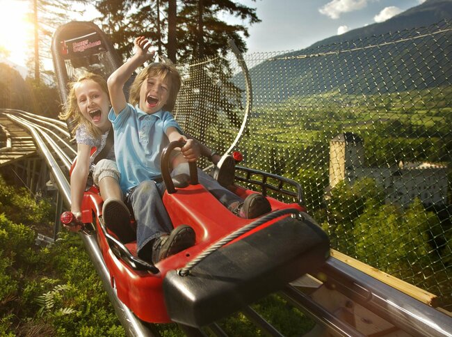 Zwei Kinder fahren lachend mit der roten Sommerrodelbahn in Lienz in Osttirol. Im Hintergrund steht Schloss Bruck.