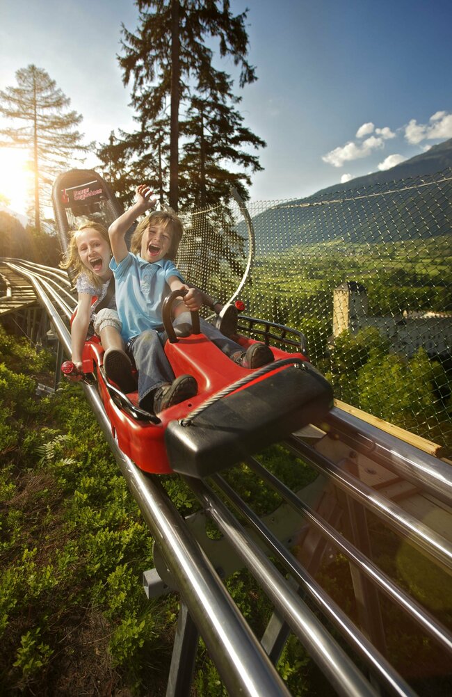 Zwei Kinder fahren lachend mit der roten Sommerrodelbahn in Lienz in Osttirol. Im Hintergrund steht Schloss Bruck.