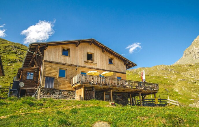 Das Bild zeigt die Eisseehütte in hochalpiner Lage auf einer grünen Bergwiese. Das Gebäude besteht aus einer Holzfassade mit mehreren Fenstern und einer erhöhten Terrasse mit Sonnenschirmen und einer Fahne. Vor der Hütte befinden sich ein Zaun und ein Wanderweg, während die umliegenden Berghänge bis an das Gebäude heranreichen.