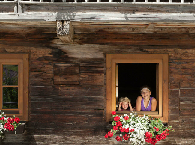 Zwei Kinder schauen aus dem Fenster eines Bauernhofs. An den Balkonen sind weiße und rote Blumen.