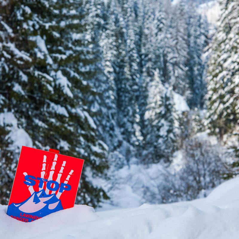 Eine rot, weiß, blaue Lawinengefahrtafel in einem frisch verschneitem Wald.