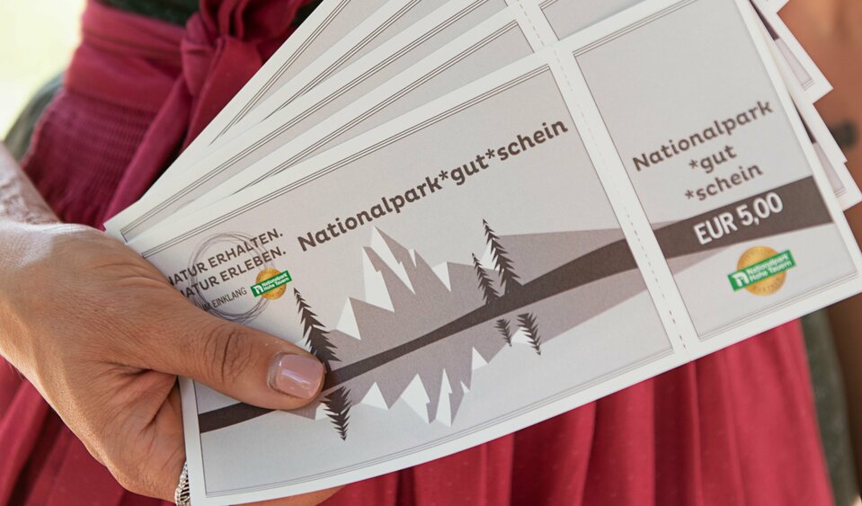 Frau im Dirndl zeigt mehrere Nationalpark Gutscheine im Wert von 5 Euro.