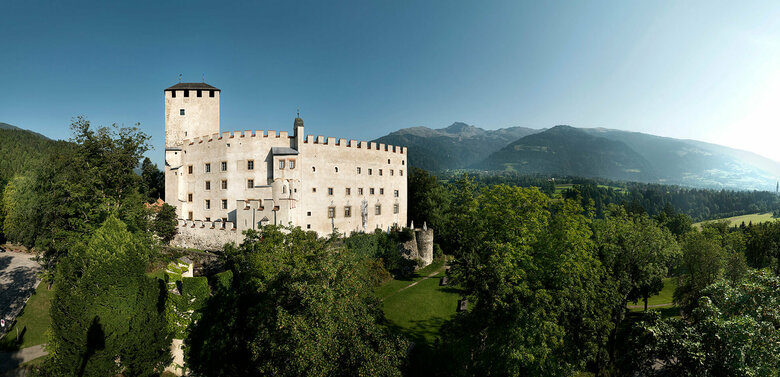 Das steinerne Schloss Bruck in Lienz umgeben von grüner Vegetation, an einem sonnigen Tag, mit Bergkulisse im Hintergrund.
