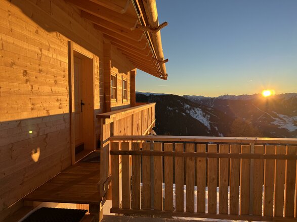 Ein Holzhaus mit Balkon, das in die Berge blickt. Die Sonne geht hinter den Gipfeln auf und sorgt für eine wunderschöne Stimmung.