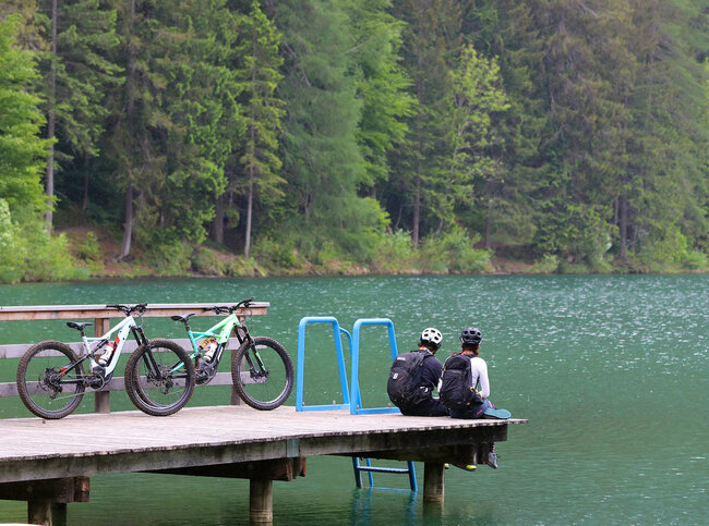 Ein Pärchen sitzt auf dem Holzsteg am Tristacher See und blickt aufs Wasser. Hinter ihenen stehen ihre E-Bikes.