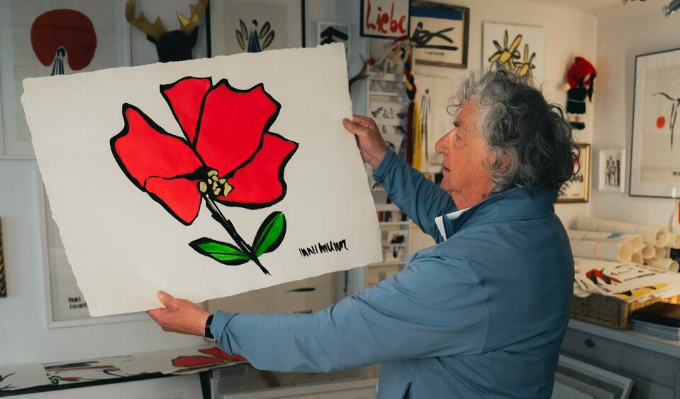Hans Salcher hält eines seiner Werke, auf dem er die Lienz Rose gemalt hat. Im Hintergrund sieht man weitere seiner Malereien. Auf dem Tisch neben ihm ist ein Bild mit dem Schriftzug Sommerfest Lienz für das Logo. 