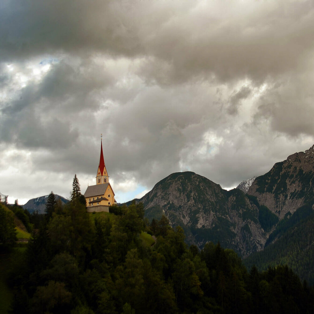 Kirche St. Justina inmitten eines Hanges umgeben von Bäumen und den Lienzer Dolomiten im Hintergrund.