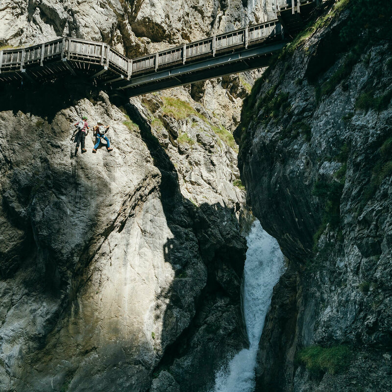 Zwei Personen auf dem Klettersteig in der Galitzenklamm. Sie befinden sich unterhalb des hölzernen Weges und neben einem Wasserfall.