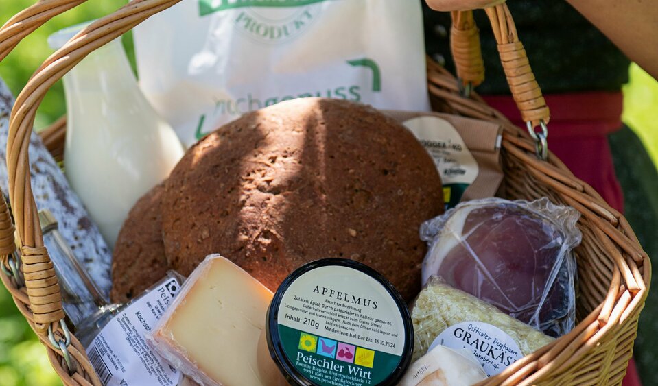 Einen Frau in Dirndl hält einen Holzkorb, gefüllt mit zahlreichen regionalen Produkten in der Hand.