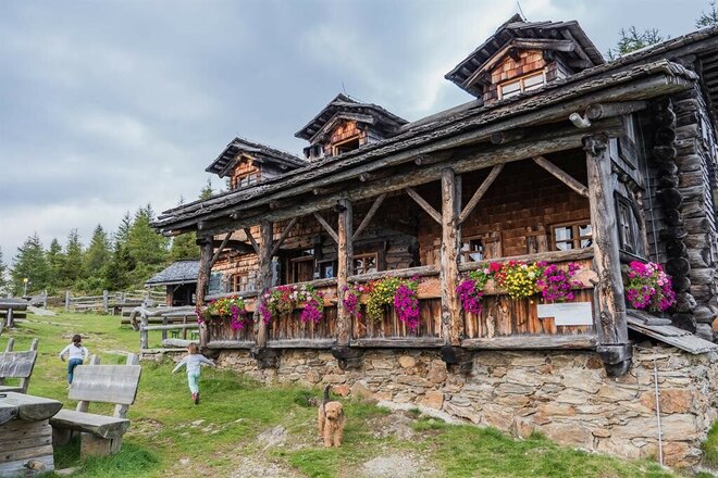 Eine traditionelle alpine Schutzhütte aus dunklem Altholz auf einem Steinsockel. Die überdachte Holzveranda ist mit leuchtend pinken und gelben Blumenkästen dekoriert. Im Vordergrund rennen zwei kleine Mädchen über die grüne Wiese, während ein struppiger, brauner Hund (Terrier-Mischling) in die Kamera blickt. Im Hintergrund sind Nadelbäume unter einem bewölkten Himmel zu sehen.