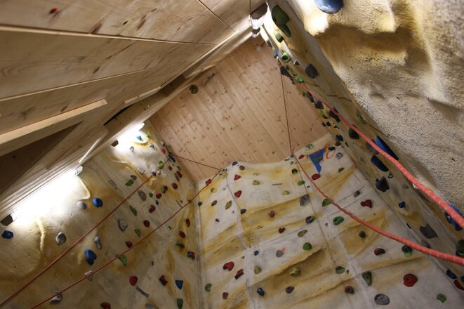 Das Bild zeigt den Kletterturm in der Sajathütte. Die Kletterwände reichen über mehrere Ebenen bis unter die Holzdecke und sind mit zahlreichen bunten Klettergriffen ausgestattet. Entlang der Wände verlaufen mehrere Kletterseile, die von oben herabhängen.