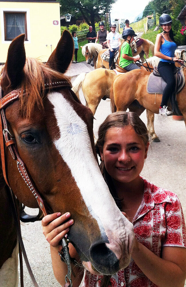 Ein Mädchen steht neben einem Pferd mit Trense auf einem Reitbauernhof. Im Hintergrund sind weitere Personen auf Pferden.