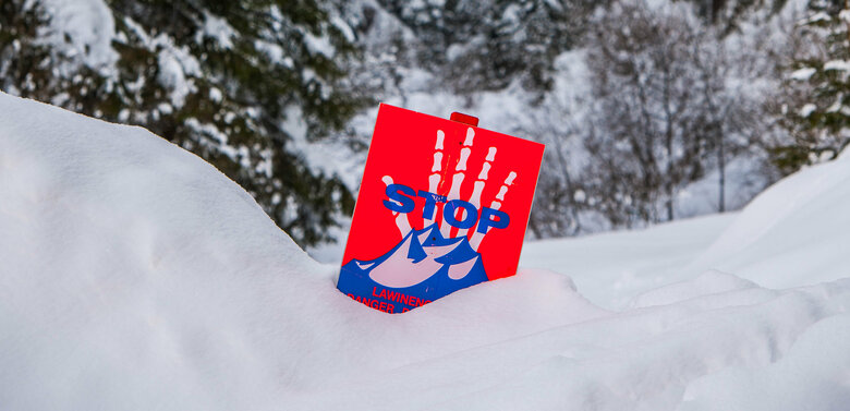Ein rot-weiß-blaues Warnschild steckt im tiefen Schnee und warnt vor Lawinen.