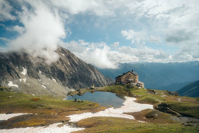 Die Sudetendeutsche Hütte steht auf einem Bergsattel inmitten einer hochalpinen Landschaft. Vor der Hütte liegt ein kleiner Bergsee, in dem sich Himmel und Berge spiegeln. Rundherum sind felsige Hänge, Schneefelder und weite Bergpanoramen zu sehen.