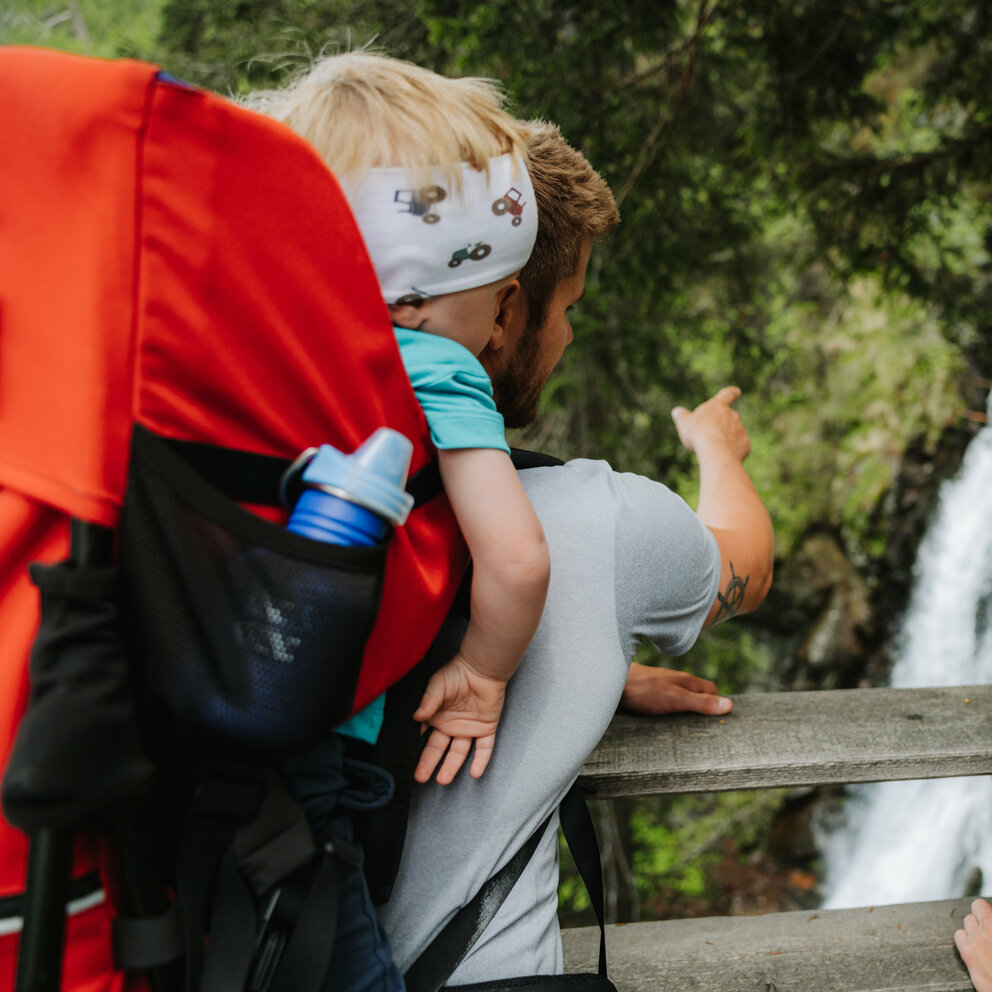 Ein Mann mit einem kleinen Kind auf dem Rücken zeigt einem Mädchen den Wasserfall im Kristeinertal.