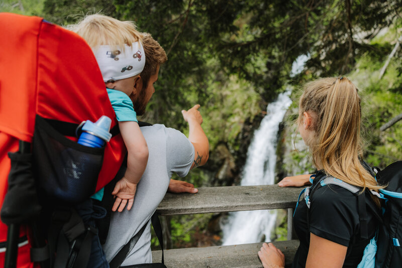 Ein Mann mit einem kleinen Kind auf dem Rücken zeigt einem Mädchen den Wasserfall im Kristeinertal.