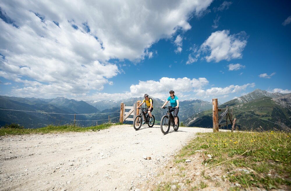 Ein Paar auf E-Bikes auf der MTB-Route 147 am Klaunzerberg. Die zwei Personen fahren auf einem breiten Weg durch ein geöffnetes Weidegatter. Im Hintergrund erstreckt sich ein weitläufiges Bergpanorama.