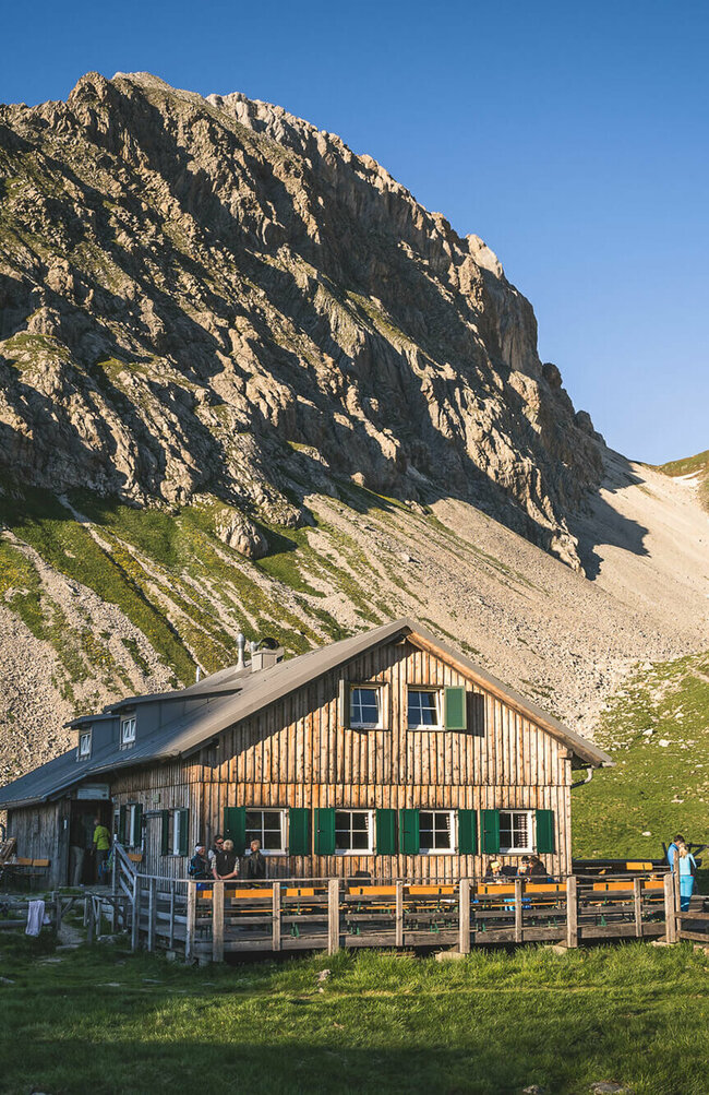 Ein rustikales Berghaus aus Holz mit grünen Fensterläden steht in einer sonnigen, alpinen Wiesenlandschaft. Im Hintergrund ragen steile, felsige Berggipfel auf, teils mit Schneefeldern bedeckt. Vor dem Haus sitzen und stehen einige Menschen, eine Holzterrasse mit Bänken lädt zum Verweilen ein