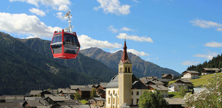 Sommergondel der Obertilliach Bergbahn fährt am Tal und der Kirche Obertilliachs vorbei