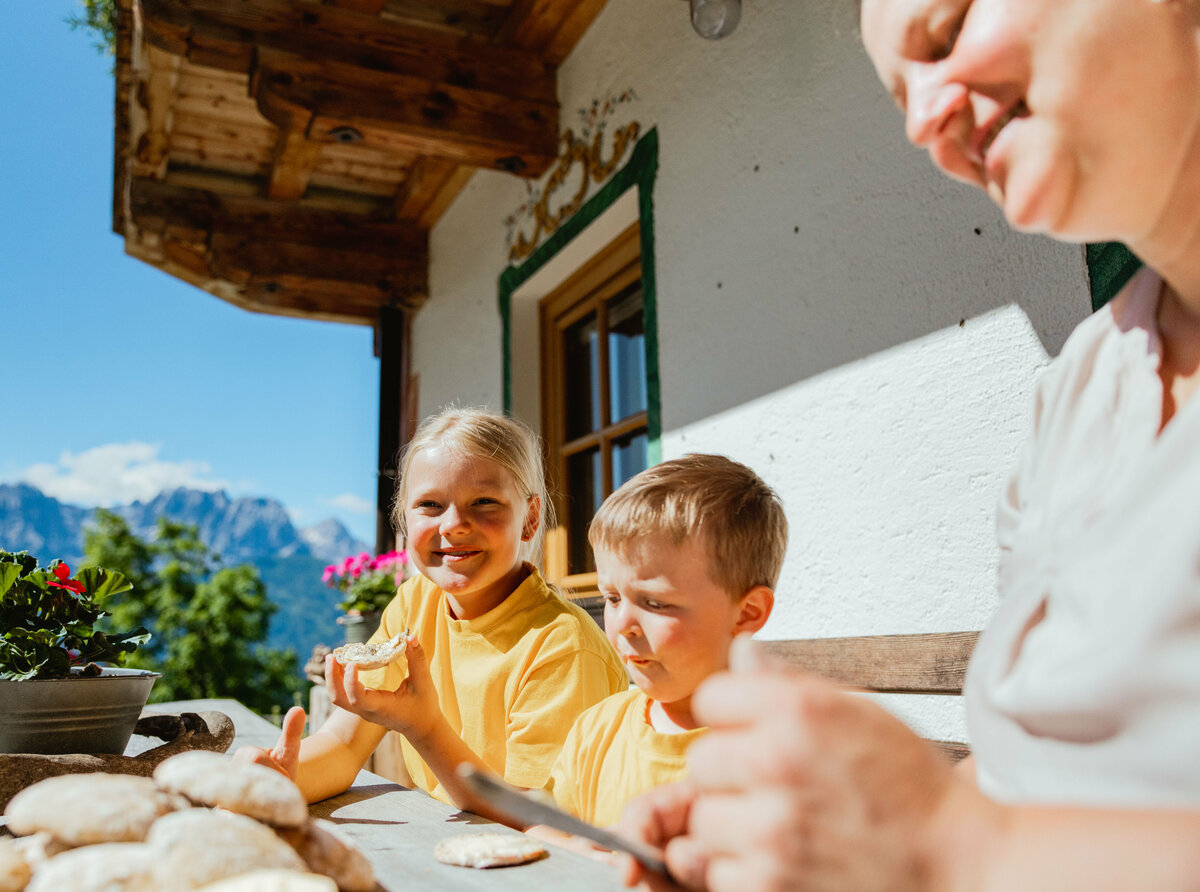 Eine Frau und zwei Kinder mit gelben T-Shirts sitzen vor einem Bauernhaus auf einer Bank und verköstigen frisches Brot.