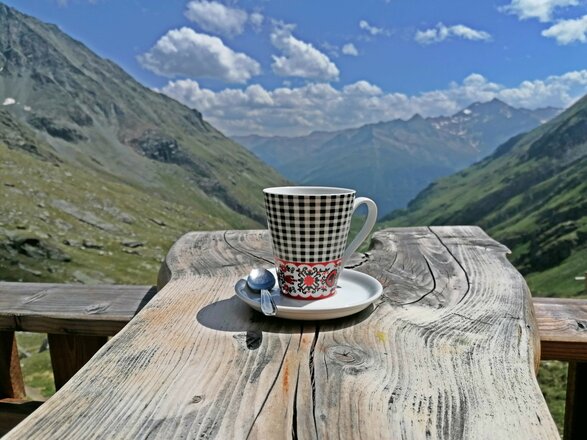 Das Bild zeigt den Ausblick von der Terrasse der Eisseehütte. Im Vordergrund stehen eine Tasse Kaffee auf einem Unterteller sowie einige Beeren auf einem rustikalen Holztisch. Dahinter öffnet sich der Blick in ein weites Alpental mit grünen Berghängen und zahlreichen Gipfeln unter einem blauen Himmel mit einzelnen Wolken.