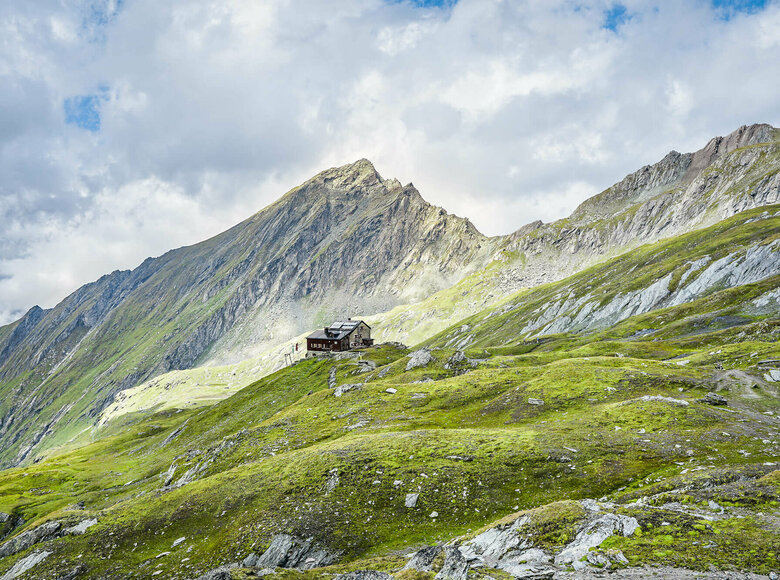 Blick auf die Sudentendeutsch Huette am Adlerweg in Osttirol, Etappe 7. Umgeben von einer felsigen Berglandschaft, teilweise grün bewachsen.