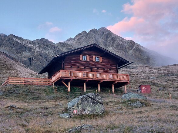 Holzhütte mit Terrasse vor einem felsigen Berggipfel in der Abenddämmerung.