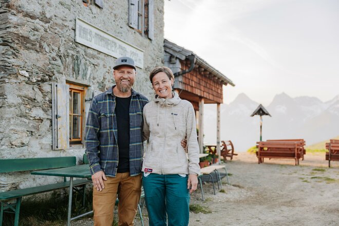 Das Bild zeigt die Pächter des Kals-Matreier-Törl Hauses vor der Hütte. Die beiden Personen stehen neben der steinernen Außenwand des Gebäudes, während im Hintergrund Sitzbänke, die Terrasse und die umliegende Berglandschaft zu sehen sind. Die Aufnahme vermittelt einen Eindruck von den Gastgebern der Berghütte in ihrer alpinen Umgebung.