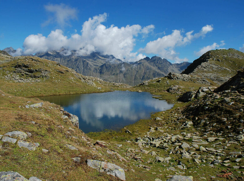 Einer der Neualpenseen am Lienzer Zettersfeld, im Vordergrund der klare Bergsee, im Hintergrund ein mächtiges Bergmassiv unter blauem Himmel.