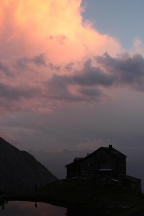 Die Sudetendeutsche Hütte ist in der Dämmerung als dunkle Silhouette vor einem farbenfrohen Himmel zu sehen. Rosa und orange gefärbte Wolken prägen die Stimmung über den umliegenden Bergen. Im Vordergrund sind Teile des Geländes und ein kleiner Bergsee erkennbar.