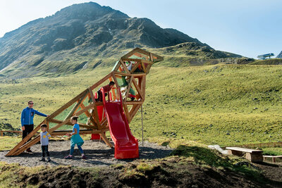 Zu sehen sind glückliche Kinder auf einem Spielgerät, welches einem überdimensional-großen Vogel ähnlich sieht, des Wassermythos Ochsenlacke im Skizentrum St. Jakob i. D.. Das sonnige Wetter lässt die umliegende Umgebung und die Berge im Hintergrund in einem warmen Licht erstrahlen.