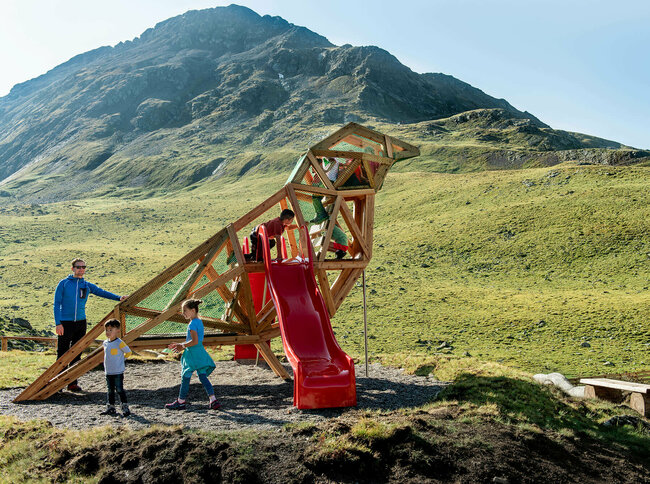 Zu sehen sind glückliche Kinder auf einem Spielgerät, welches einem überdimensional-großen Vogel ähnlich sieht, des Wassermythos Ochsenlacke im Skizentrum St. Jakob i. D.. Das sonnige Wetter lässt die umliegende Umgebung und die Berge im Hintergrund in einem warmen Licht erstrahlen.