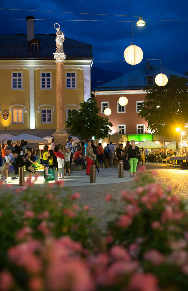 Aufnahme von mehreren Personen beim Moonlight Shoppen in Lienz. Es ist Nacht, die Straßenbeleuchtung ist eingeschalten und das Foto wurde vor einem Blumenbeet gemacht.