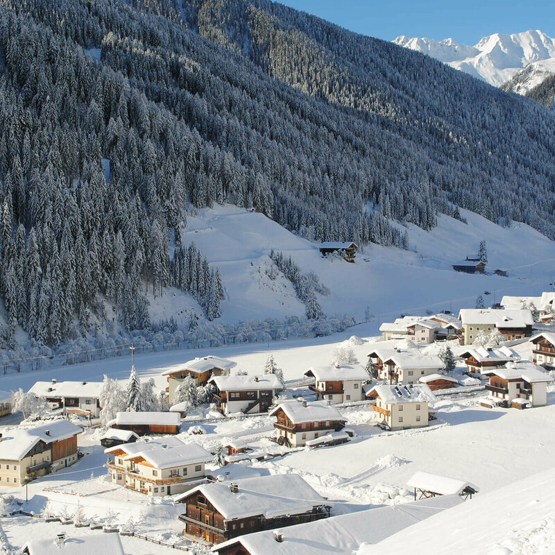 Blick über das verschneite Bergdorf Innervillgraten, hinter dem sich die bewaldete Bergwand empor streckt, an einem sonnigen Wintertag.