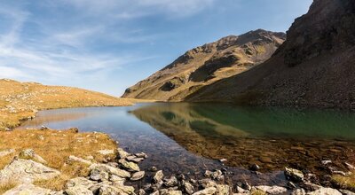Der Blick auf den Mond- & Schwarzsee in Hopfgarten i. D.. Das klare Wasser spiegelt den blauen Himmel und die umgebende felsige Bergwelt in sich. Das Ufer ist bräunlich gefärbt, was vermuten lässt, dass diese Aufnahme im Spätherbst gemacht wurde.