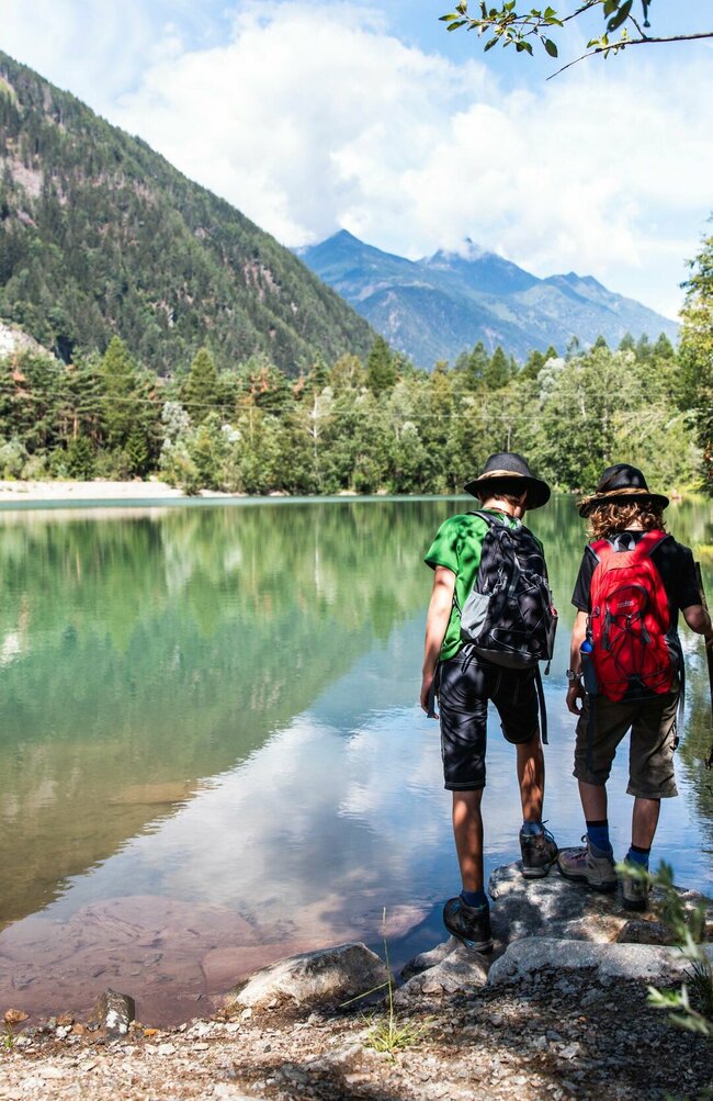 Zwei Personen mit Wanderausrüstung und Hüten stehen an einem See und blicken in die Ferne.