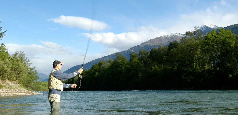 Ein Mann steht in einem Fluss und wirft seine Angel aus zum Fliegen fischen.