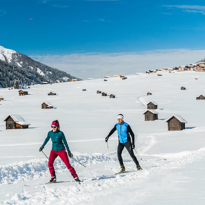 Zwei Langläufer:innen skaten auf der Loipe in Obertilliach bei herrlichem Winterwetter durch die Tilliacher Feldern, die übersäht mit zahlreichen kleinen Heustadeln und zwei kleinen Kapellen sind. Dahinter erhebt sich das Dorf.