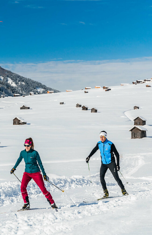 Zwei Wintersportler laufen auf ihren Langlaufskiern auf der Loipe in den Obertilliacher Feldern mit den zahlreichen kleinen Hütten und dem Dorf im Hintergrund