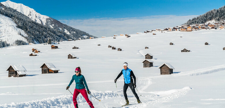 Zwei Wintersportler laufen auf ihren Langlaufskiern auf der Loipe in den Obertilliacher Feldern mit den zahlreichen kleinen Hütten und dem Dorf im Hintergrund