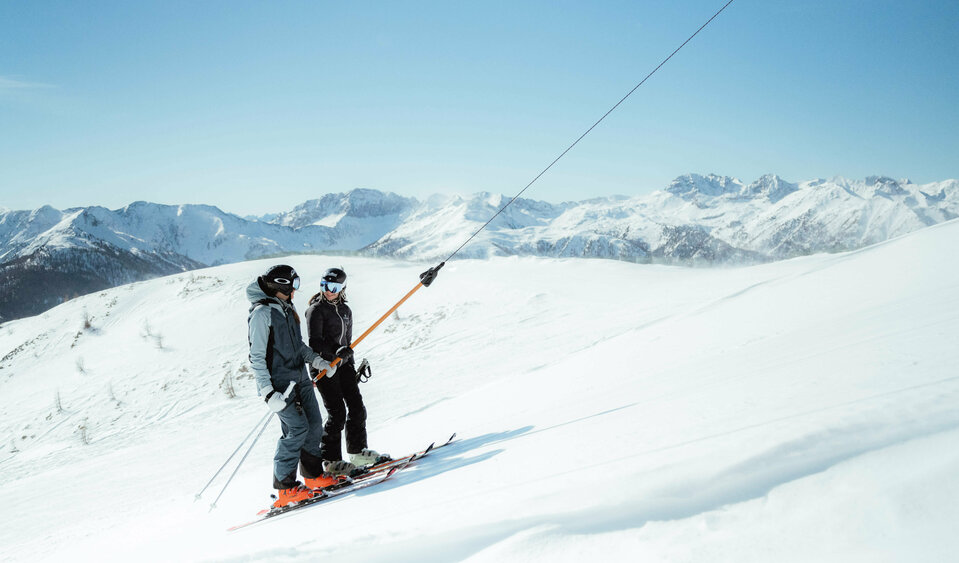 Zwei Skifahrer lassen sich vom Schlepplift im Skigebiet Golzentipp in Obertilliach hoch auf den Berg ziehen mit herrlichem Winterpanorama und schönem Wetter.