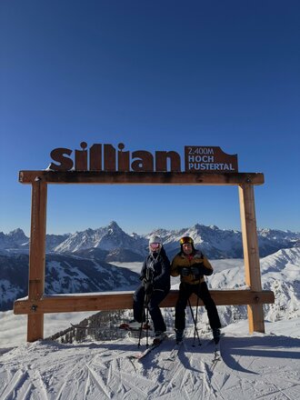 Zwei Skifahrer stehen vor dem Schild "Sillian 2.400m Hochpustertal" in den Bergen. Im Hintergrund sind schneebedeckte Gipfel und ein klarer blauer Himmel zu sehen.