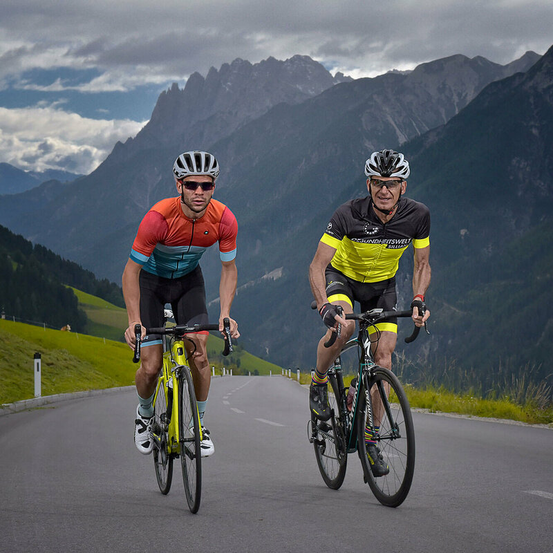 Zwei Rennradfahrer auf der Pustertalerhoehenstrasse mit bedrohlicher Berguklisse und dunklen Wolken im Hintergrund.
