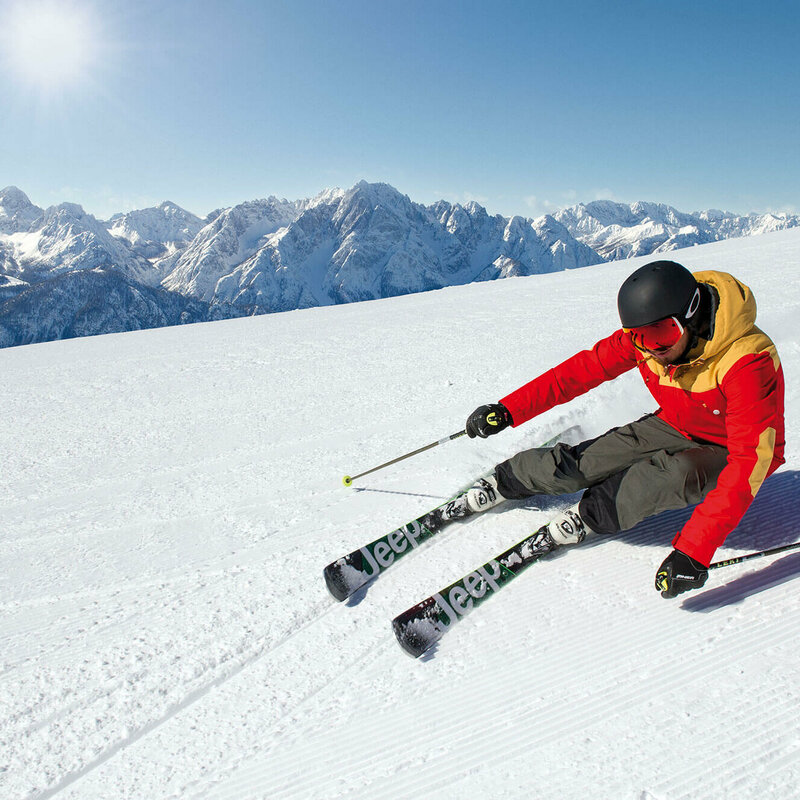 Skifahrer mit rot-gelber Jacke und schwarzem Helm beim Terrassenskilauf am Zettersfeld Skigebiet. Die Sonne strahlt und der Himmel ist blau.